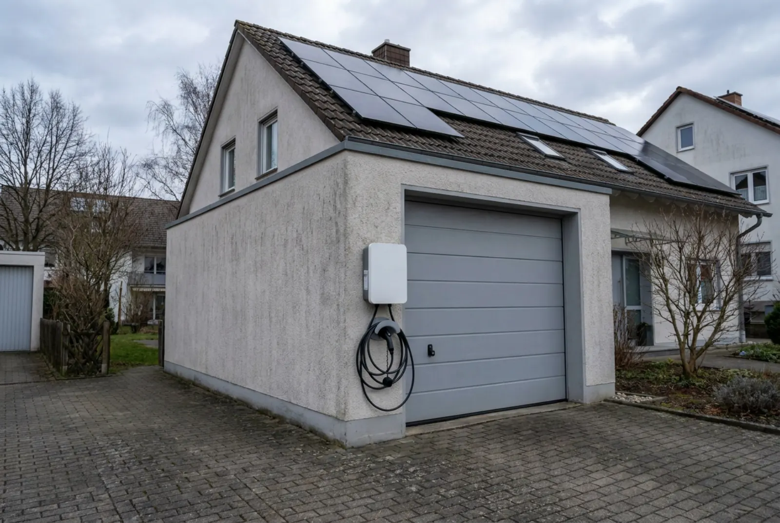 Weiße Wallbox mit aufgerolltem Ladekabel an der Garage neben dem Tor, auf dem Hausdach Solarmodule