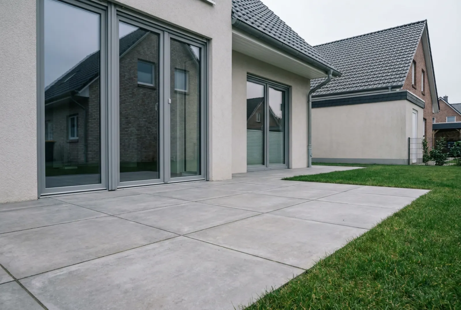 Terrasse mit frisch verlegten großformatigen Platten in Betonoptik vor bodentiefen Fenstern, daneben Rasen