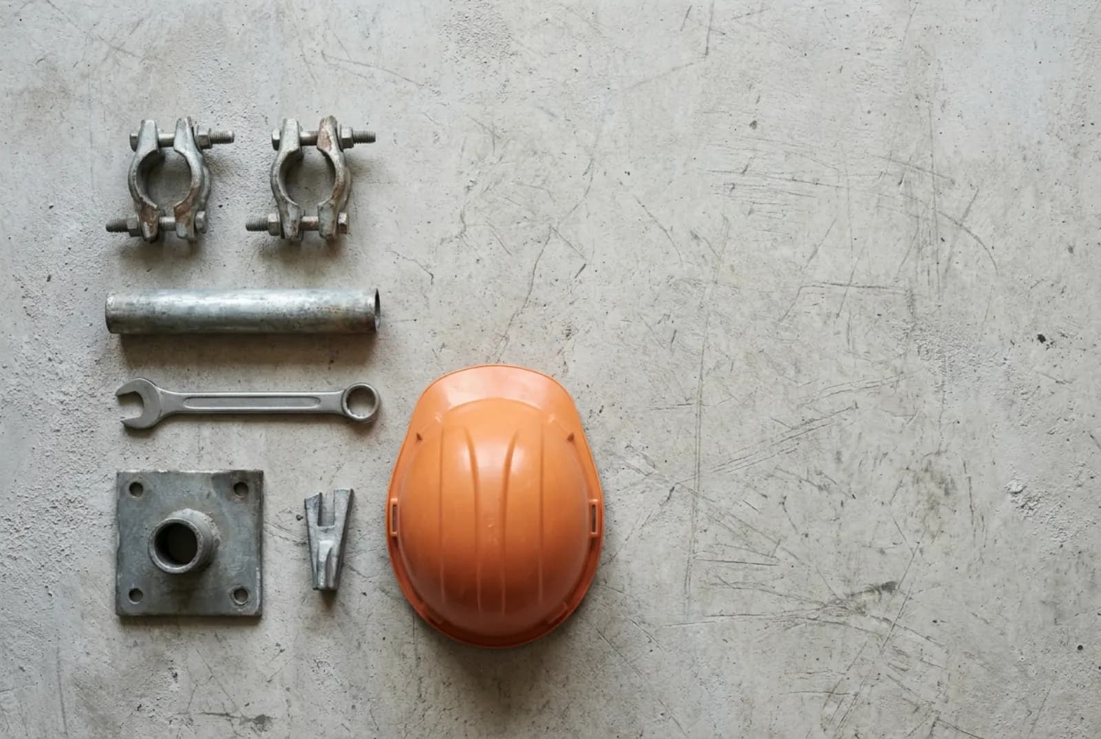 Gerüstkupplungen, Stahlrohr, Ringmaulschlüssel, Fußplatte, Keilverbinder und ein oranger Schutzhelm ohne Aufdruck auf einer Betonwerkbank
