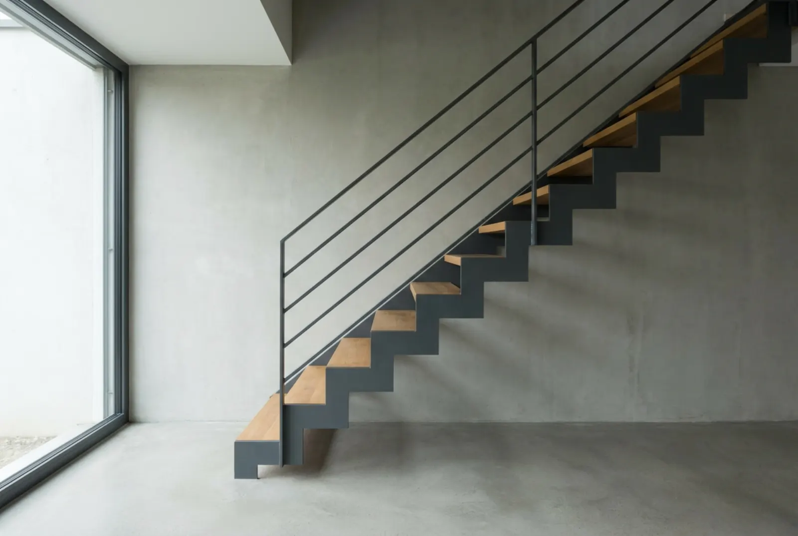 Stahltreppe mit Flachstahlgeländer und Eichenstufen vor einer Sichtbetonwand, Licht von einem großen Fenster