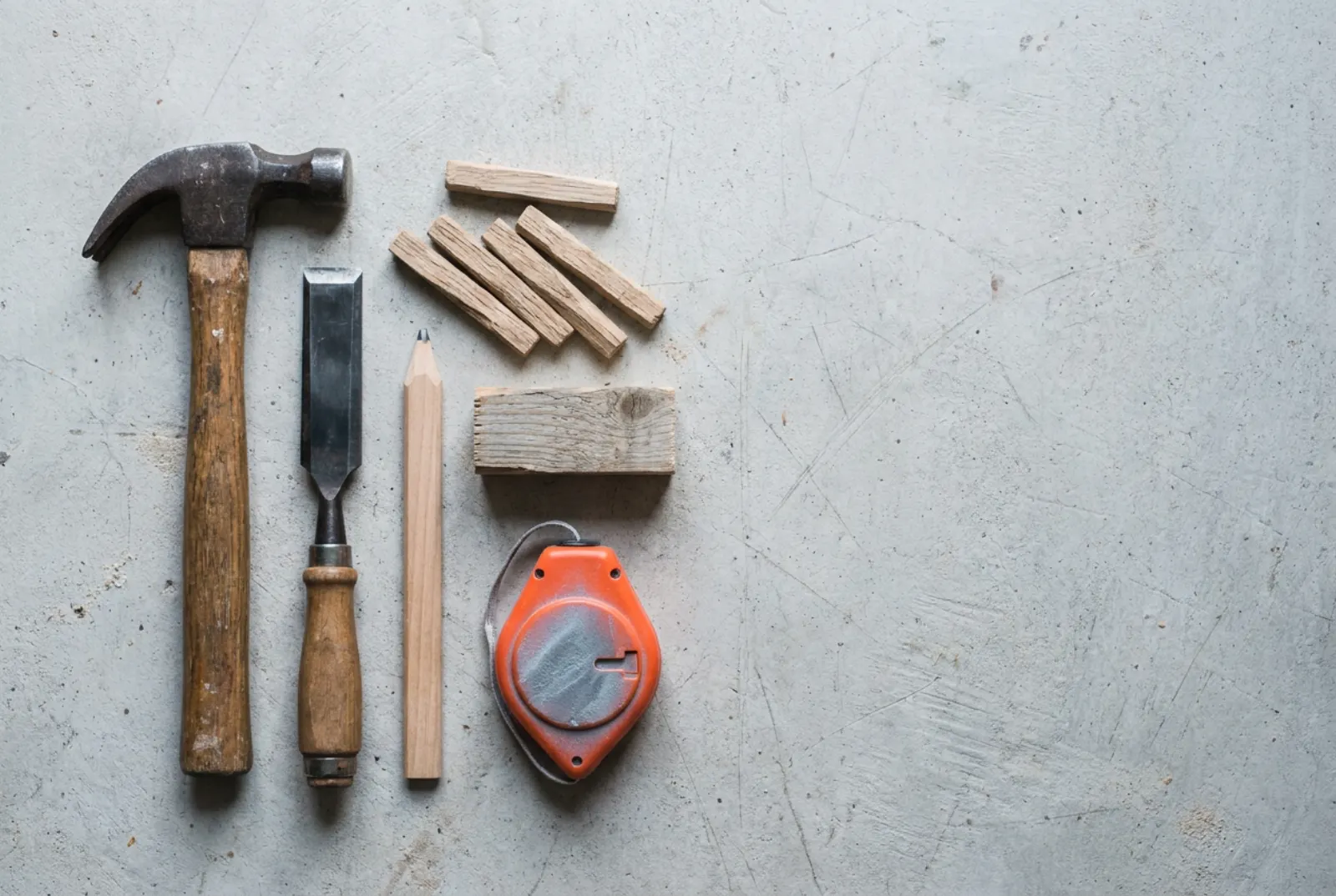 Hammer, Stemmeisen, Bleistift, Holznägel aus Eiche, Fichtenabschnitt und Schlagschnur mit orangem Gehäuse auf einer Betonwerkbank