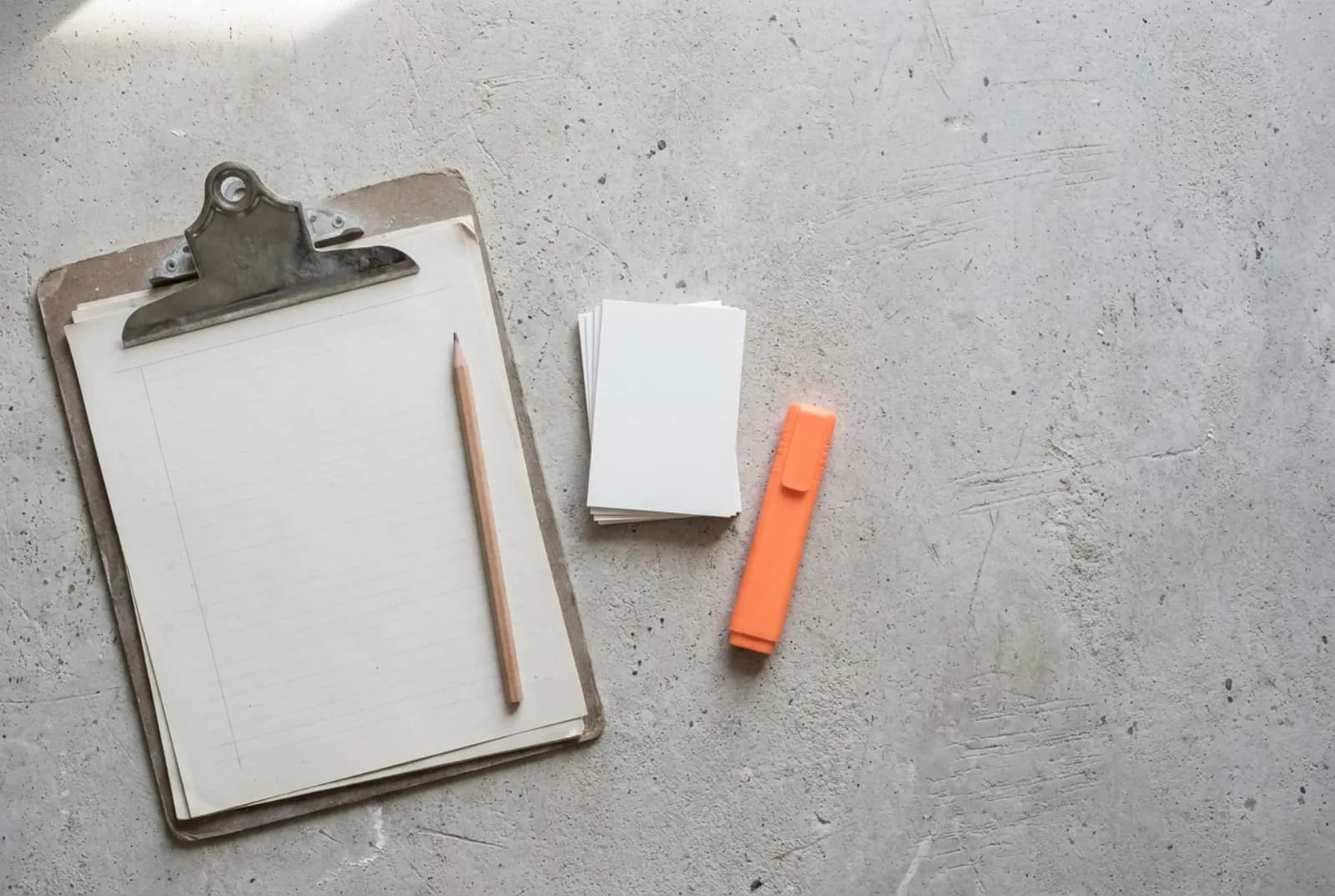 Klemmbrett mit leeren linierten Blättern, Bleistift, unbedruckte Karten und ein orangefarbener Textmarker auf einer Betonwerkbank