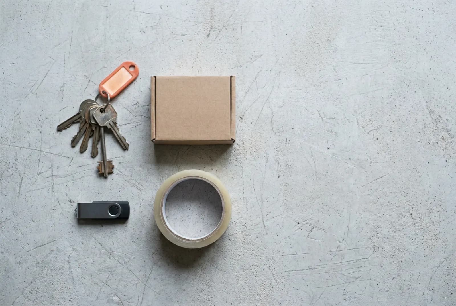 Schlüsselbund mit orangem Anhänger, kleiner Umzugskarton, USB-Stick und Packband auf einer Betonwerkbank