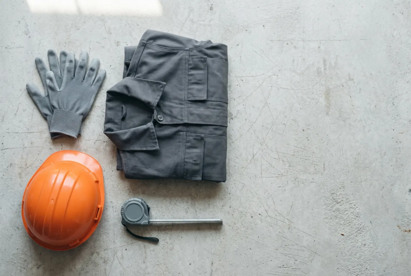 Neue Arbeitshandschuhe, eine gefaltete Arbeitsjacke, ein oranger Schutzhelm und ein Maßband auf einer Betonwerkbank